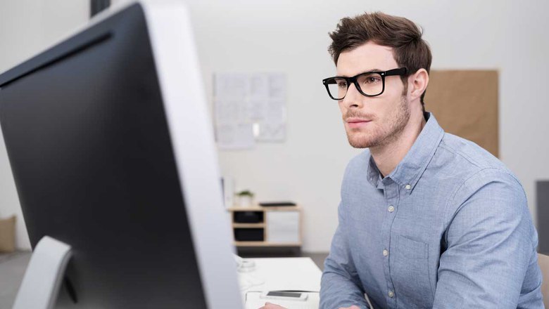 Специалист по SEO-продвижению в серой рубашке и очках перед экраном компьютера в офисе.